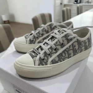Gråvita sneakers från Dior - Säljer ett par snygga sneakers från Dior med grått och vitt mönster och tydlig logga. Skorna har vita snören, rund tå och platt sula. Perfekta för dig som gillar stilrena och trendiga skor.                  [Box, dust bags och äkthetsbevis finns]