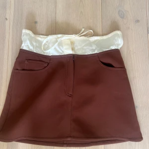 Brun minikjol med satinmidja från Acne Studios - Säljer en brun minikjol från Acne Studios med unik vit satinmidja och knytband. Kjolen har fickor framtill och en snygg, stilren design. Kjolen är i nyskick. Strlk S.