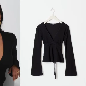 Svart omlottopp med knytband - Svart omlottopp med långa ärmar och v-ringning. Toppen har knytband i midjan och lätt utsvängda ärmar. Perfekt för en trendig och stilren look.