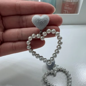Hjärtformade pärlörhängen med grått hjärta - Superfina örhängen i form av hjärtan, dekorerade med vita pärlor och ett mjukt grått tygklätt hjärta upptill. Perfekta för att ge en söt och trendig touch till din outfit.