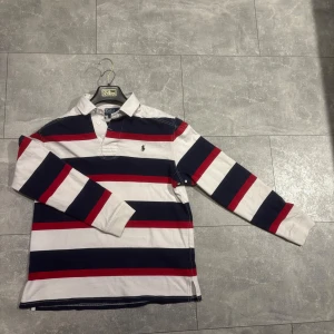 Randig långärmad pikétröja från Polo Ralph Lauren - Säljer en klassisk långärmad pikétröja från Polo Ralph Lauren med breda ränder i vitt, marinblått och rött. Tröjan har krage, knappar vid halsen och den ikoniska broderade loggan på bröstet.