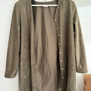 Beige kappa/kofta från Comfort Fashion - Säljer en beige kappa/kofta från Comfort Fashion i storlek 42/44. Jackan har långa ärmar, dekorativa knappar framtill och fina utskurna detaljer längst ner. Materialet känns mjukt och har en mockaliknande yta.