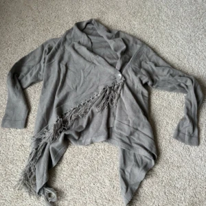 Grå kofta med fransar från Soyaconcept - Säljer en grå kofta från Soyaconcept med långa ärmar, asymmetrisk nederkant och fransdetaljer framtill. Koftan har en knappstängning och är tillverkad i mjukt stickat material. Perfekt för lager-på-lager-stil.