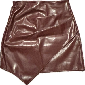 Röd skinnkjol med omlott-detalj - Säljer en röd kort skinnkjol med omlott-detalj framtill. Kjolen har en asymmetrisk nederkant och en glansig finish som ger en cool look. Perfekt för dig som vill sticka ut lite extra.