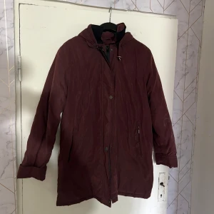 Vinröd parkajacka från Best Comfort - Säljer en vinröd parkajacka från Best Comfort med huva och dragkedja framtill. Jackan har långa ärmar, knappar och två fickor på sidorna. Perfekt för kyligare dagar och har en klassisk, rak passform.
