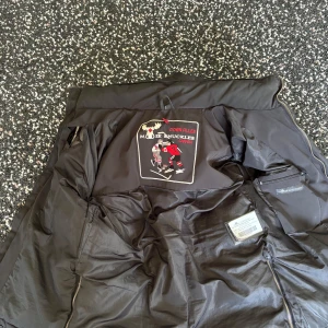 Svart dunjacka med fuskpäls från Moose Knuckles - Säljer en svart dunjacka från Moose Knuckles med avtagbar huva och fuskpäls. Jackan har flera fickor med dragkedja och knappar samt logga på ärmen. Perfekt för kalla dagar.