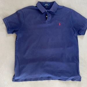 Blå pikétröja från Polo Ralph Lauren - Säljer en klassisk blå pikétröja från Polo Ralph Lauren i storlek XL. Tröjan har korta ärmar, krage med knappar och den ikoniska röda broderade loggan på bröstet. Perfekt för en stilren och avslappnad look.