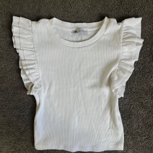 Vit ribbad topp med volangärmar från Zara - Supersöt vit ribbad topp från Zara med volangdetaljer på axlarna. Perfekt för att piffa upp en enkel outfit. Mjuk och stretchig, passar till många olika stilar.