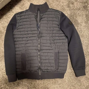 Mörkblå cardigan  - Säljer en mörkblå cardiganmed quiltad framsida och rygg samt släta ärmar. Jackan har dragkedja framtill, ribbade muddar och ståkrage. Perfekt för dig som vill ha en stilren och sportig look.