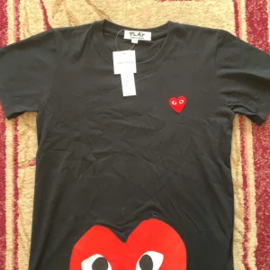 Svart t-shirt med hjärtdetalj från Comme des Garçons Play - Svart t-shirt från Comme des Garçons Play med ikoniskt rött hjärta med ögon på bröstet och ett större hjärta nedtill. Klassisk rund hals och korta ärmar. Perfekt för dig som gillar streetwear och vill sticka ut med en cool detalj.