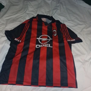 AC Milan fotbollströja med Opel sponsor - Säljer en klassisk AC Milan fotbollströja från Adidas med röd-svart randigt mönster och Opel som sponsor på bröstet. Tröjan har korta ärmar och klubbmärket på vänster bröst. Perfekt för dig som älskar italiensk fotboll!