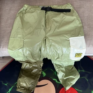 Ljusgröna täckbyxor från 66°North x Ganni - Säljer ett par ljusgröna täckbyxor från 66°North x Ganni med justerbart bälte i midjan och flera fickor. Byxorna har en vit detalj på ena benet och elastiska benslut. Perfekta för kalla dagar och outdoor-aktiviteter.