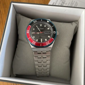 Timex automatisk klocka med stållänk - Säljer en snygg automatisk Timex-klocka med stållänk. Urtavlan är svart med dag- och datumvisning, och vridringen har röd och blå färg. Klockan levereras i originalask. Perfekt för dig som gillar klassisk stil med en sportig touch. Nu pris ligger runt 2000