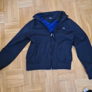 Svart vindjacka från Fred Perry - Snygg svart vindjacka från Fred Perry med dragkedja framtill och diskret logga på bröstet. Jackan har hög krage, två fickor och blått meshfoder på insidan. Perfekt för en stilren look.