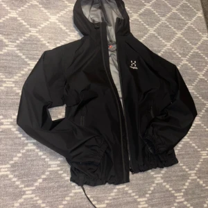 Svart vindjacka från Haglöfs med huva - Säljer en svart vindjacka från Haglöfs i storlek M. Jackan har huva, dragkedja framtill och elastiska muddar vid ärmslut och midja. Perfekt för blåsiga dagar och outdoor-aktiviteter. Innerfodret är i mesh för extra ventilation.