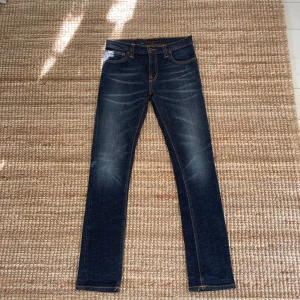Mörkblå jeans från Nudie Jeans - Säljer ett par klassiska mörkblå jeans från Nudie Jeans med orangea sömmar och signaturbroderi på bakfickan. Jeansen har fem fickor och raka ben. Perfekta för en stilren vardagslook.