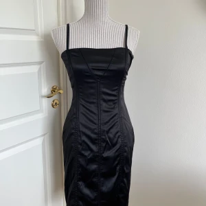 Svart axelbandslös klänning med snörning - Snygg svart klänning i satin med axelbandslös design och snörning i ryggen. Perfekt för festliga tillfällen. Klänningen har en tight passform som framhäver figuren. 