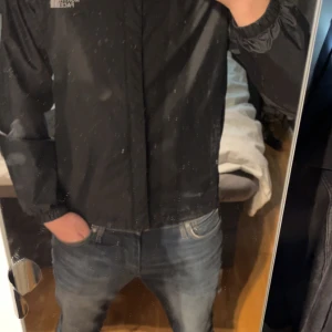 Svart vindjacka från The North Face - Säljer en svart vindjacka från The North Face med huva och dragkedja. Jackan är lätt och har en enkel, stilren design med logga på bröstet och ryggen. Perfekt för blåsiga dagar och enkel att packa ner i väskan.OBS har sytt själv i armvecken, men den fungerar exakt som vanligt !!!! Säljer för att den är för liten. Pris kan diskuteras!