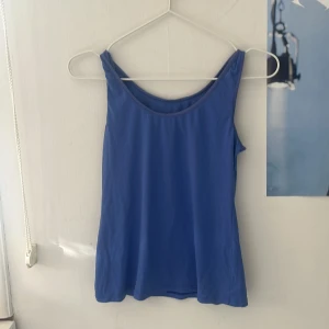 Blå topp - Säljer en enkel blå linnetopp med breda axelband och rund halsringning. Toppen är ärmlös och har en slät yta, perfekt för varma dagar eller som lager under andra plagg.