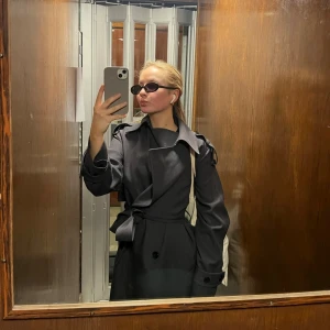 Mörkgrå trenchcoat med bälte - Säljer en stilren mörkgrå trenchcoat med klassisk krage och knytskärp i midjan. Jackan har dubbelknäppning, axelklaffar och långa ärmar med knappar vid ärmslut. Perfekt för dig som vill ha en tidlös och trendig look.