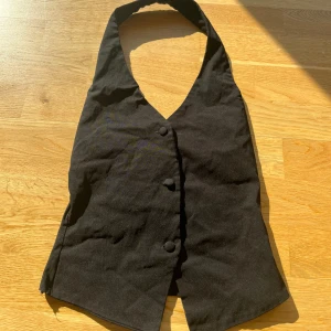 Svart halterneck väst från Boohoo - Säljer en svart halterneck väst från Boohoo med knappar framtill. Västen har en v-ringad skärning och är figurnära i modellen. Perfekt till fest.