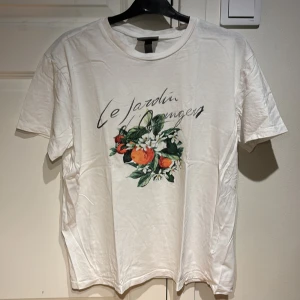 Vit t-shirt H&M - Vit t-shirt från H&M med tryck framtill som föreställer apelsiner och blommor samt texten 'Le Jardin Oranger'. Klassisk rund halsringning och korta ärmar. Perfekt för en fräsch och somrig look.