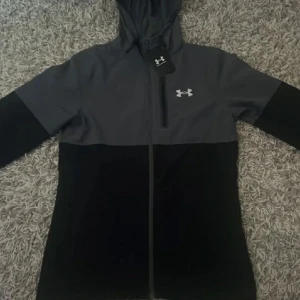 Svart och mörkgrå hoodiejacka från Under Armour - Snygg svart och mörkgrå hoodiejacka från Under Armour med dragkedja framtill och logga på bröstet. Jackan har huva och långa ärmar, perfekt för träning eller vardag. Stilren design med tvådelad färg.