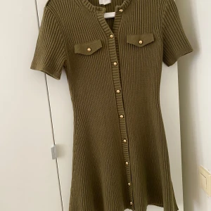 Olivgrön ribbad klänning från H&M - Säljer en olivgrön ribbad klänning från H&M med korta ärmar och dekorativa knappar framtill. Klänningen har två bröstfickor med knappdetaljer och en figurnära passform som ger en snygg siluett.