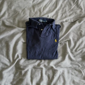 Mörkblå pikétröja från Ralph Lauren - Säljer en stilren mörkblå pikétröja från Ralph Lauren med klassisk krage och knappar. Tröjan har en broderad gul logotyp på bröstet och är i slim fit. Perfekt för en avslappnad men ändå snygg look.