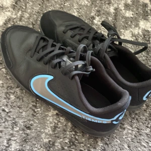 Nike Tiempo fotbollsskor med blå detaljer - Säljer ett par svarta Nike Tiempo fotbollsskor med blå och silverfärgade detaljer. Skorna har snörning och en platt sula, perfekt för inomhusspel. Snygg design med tydlig Nike-logga på sidan och hälen. Den har varit använd 1 gång men man ser inte det.