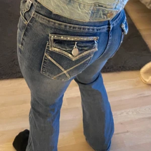 Blå bootcut jeans med detaljerade bakfickor - Säljer ett par blå bootcut jeans med snygga detaljer på bakfickorna, inklusive dekorativa sömmar och knappar. Jeansen har normal passform och klassisk femficksdesign. Perfekta för dig som vill ha ett par jeans med lite extra stil.