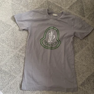 Grå t-shirt från Moncler med logga - Säljer en grå t-shirt från Moncler med stor grön och vit logga på bröstet. Klassisk rund halsringning och korta ärmar. Perfekt för dig som gillar stilrena märkesplagg. Helt ny ungefär