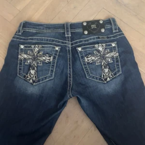 Mörkblå jeansbyxor från Miss Me med broderade bakfickor - Säljer ett par mörkblå jeans från Miss Me med snygga broderade kors på bakfickorna och dekorativa nitar. Byxorna har mid waist men sitter som low waist, bootcut-modell och dragkedja. Perfekta för dig som vill sticka ut med detaljerade jeans! Köpte för 899 för ungefär 2 veckor sen, det är för långa på mig och jag är 161. Dem är lite stora i storleken men det äe storlek W25.