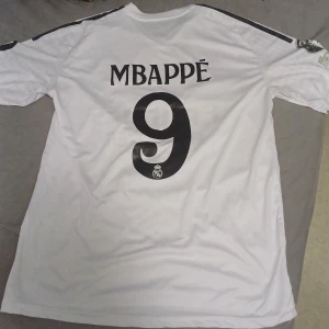 Real Madrid Mbappé 9 fotbollströja - Vit Real Madrid fotbollströja med Mbappé och nummer 9 på ryggen. Tröjan har svarta detaljer, klubbmärke på bröstet och sponsortryck framtill. Klassisk V-ringning och korta ärmar. Perfekt för real madrid fans.