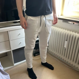 NIKE tech joggers - Säljer ett par vita mjukisbyxor från Nike med dragsko i midjan och svarta detaljer. Byxorna har normal passform, sidofickor och Nike-logga på vänster ben. Digital kvitto finns. 191 cm lång på bilden, passar perfekt.