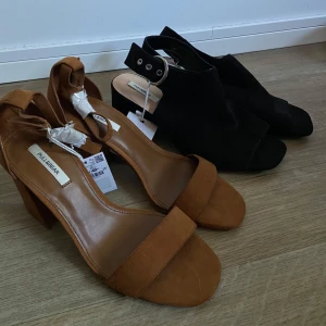 Två par klackskor från Pull&Bear - Säljer två par snygga klackskor från Pull&Bear. Ett par är bruna med öppen tå och bred klack, det andra paret är svarta med öppen tå och spänne vid ankeln. Bägge är storlek 40 och med prislapp kvar. 