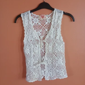 Vit virkad väst från KappAhl - Superfin bohemisk virkad väst från KappAhl med blommigt mönster och två knyten framtill. Har en liten brunare fläck på ena sidan - se bild. Väldigt liten i storlek och passar xs 🌸