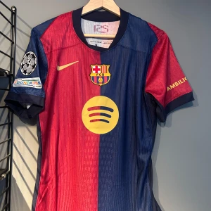 FC Barcelona matchtröja Raphinha 11 - Säljer en FC Barcelona fotbollströja med Raphinha 11 på ryggen. Tröjan är kortärmad, har röd och blå färg med klubbmärke och sponsorlogga på bröstet. Champions League-märke på ärmen. Perfekt för fans av Barca! Helt ny med tagg en replika