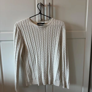 Vit kabelstickad tröja från Ralph Lauren - Säljer en klassisk vit kabelstickad tröja från Lauren Ralph Lauren. Tröjan har rund halsringning, långa ärmar och ribbade muddar vid ärmslut och nederkant. Perfekt för lager-på-lager och stilrena outfits.