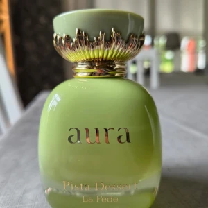 Aura Pista Dessert La Fede parfym - Doften har toppnoter av pistage, bergamott, rom och kardemumma, hjärtnoter av liljekonvalj, päron, jasmin, hallon och vit persika samt basnoter av sockervadd, tonkaböna, sandelträ och ceder.  Bra hållbarhet. Sprutat 10 max 15 gånger, går ej att se igenom men känns fortfarande fullt. Med kartong.