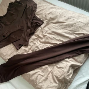 Bruna mjukisbyxor med matchande topp - Säljer ett par bruna mjukisbyxor med hög midja och tillhörande långärmad topp. Byxorna är tighta och stretchiga, perfekta för en avslappnad stil eller hemmamys. Materialet känns mjukt och följsamt mot huden.