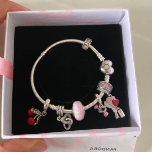 Silverfärgat armband med berlocker från Pandora - Charmigt armband i silverfärg från Pandora med flera berlocker, bland annat röda körsbär, hjärtan, rosett och en rosa pärlberlock. Perfekt accessoar för att ge en personlig touch till din stil.