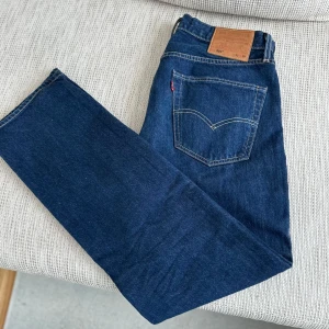 Mörkblå Levis 501 jeans - Klassiska mörkblå Levis 501 jeans med rak passform och femficksdesign. Byxorna har en tydlig Levis-lapp bak i midjan och den ikoniska röda Levis-taggen på bakfickan. Perfekta för en tidlös och avslappnad stil.