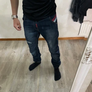 Mörkblå jeans med färgdetalj - Säljer ett par mörkblå jeans med röda och blå ränder på fickan. Jeansen har regular fit passform. Storlek S