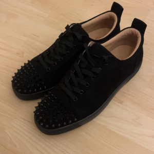  Christian Louboutin - Säljer ett par svarta sneakers från Christian Louboutin med svarta nitar på tån och klassisk röd sula. Skorna har snörning och är tillverkade i mocka med detaljer i skinn. Perfekta för dig som vill sticka ut med en edgy stil.
