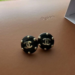 Svarta örhängen med gulddetaljer från Chanel - Säljer ett par runda svarta örhängen från Chanel med guldfärgade nitar och den klassiska CC-loggan i guld. Snygga och tidlösa accessoarer som lyfter varje outfit. Nypris: 4 675 kr – säljes till fyndpris! Original med exklusiv känsla.