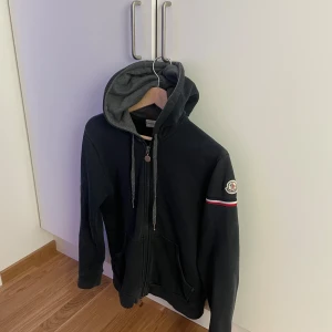 Svart hoodie från Moncler med dragkedja - Säljer en svart hoodie från Moncler med grå insida på huvan och dragkedja framtill. Tröjan har en broderad Moncler-logga på ärmen och en röd, vit och blå randdetalj. Perfekt för en avslappnad stil.