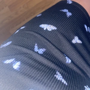 Svart kortklänning med små fjärilar - Supersöt svart kortklänning med blåa fjärilar och spetsdetaljer vid bysten. Klänningen har smala axelband och en figurnära passform. 