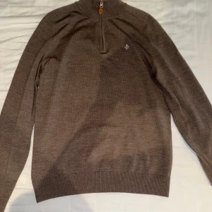 Brun grå tröja från Morris - Säljer en brun stickad tröja från Morris med halv dragkedja och broderad logga på bröstet. Tröjan har ribbade muddar och hög krage med kontrastfärg på insidan. Perfekt för lager-på-lager under kyliga dagar.