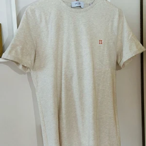 Beige t-shirt från Les Deux - Säljer en stilren beige t-shirt från Les Deux med liten broderad detalj i orange på bröstet. T-shirten har rund halsringning och korta ärmar. Perfekt för en clean och enkel look.